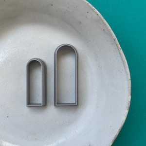 Könnte beinhalten: Zwei graue, metallene Ausstecher in Bogenform auf einem weißen Keramikteller. Der größere Ausstecher ist etwa 12,5 cm hoch und der kleinere etwa 7,5 cm hoch.