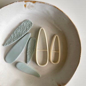 Puede incluir: Un conjunto de cinco herramientas de arcilla polimérica para manualidades. Las herramientas tienen forma de hojas y óvalos. Dos de las herramientas son de color azul claro y tienen un diseño floral. Las otras tres herramientas son blancas y tienen una superficie lisa.