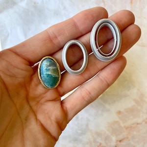 Puede incluir: Tres anillos de metal ovalados, uno con un borde dorado, y una piedra verde y blanca en el centro. Los anillos son de diferentes tamaños y se sostienen en una mano.