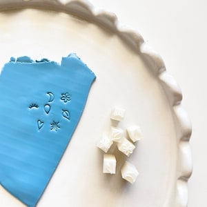 May include: A set of white polymer clay stamps with various designs, including a crescent moon, a star, a flower, a heart, an eye, a leaf, and a balloon. The stamps are on a white plate with a blue polymer clay sheet with the same designs imprinted on it.