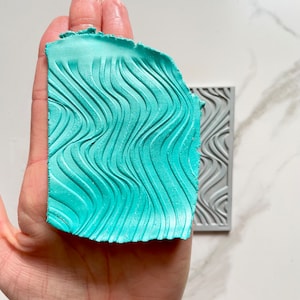 May include: A teal blue silicone mold with a wavy pattern. The mold is being held in a hand.