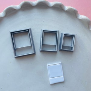 Peut inclure: Quatre emporte-pièces en plastique gris en forme de cadre photo et un emporte-pièce en plastique blanc en forme de cadre photo sur une assiette blanche à bord festonné.