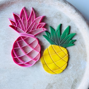May include: Two pineapple cookie cutters, one pink and one yellow with green leaves. The pink cutter is a detailed outline of a pineapple, while the yellow cutter is a solid shape.