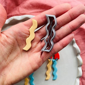 Puede incluir: Dos cortadores de galletas con forma de onda, uno de plástico gris y el otro de fondant amarillo. También hay tres piezas de fondant con forma de onda en azul, amarillo y amarillo claro.
