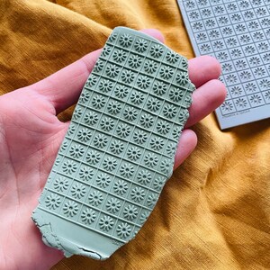 May include: A close-up of a green clay slab with a daisy pattern embossed into the surface. The clay is being held in a hand.