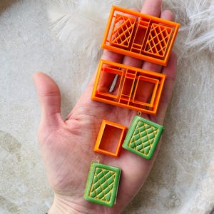 May include: Four orange plastic moulds for making polymer clay book charms. Two of the moulds are rectangular and have a grid pattern. The other two moulds are rectangular and have a simple frame design. Two green polymer clay book charms with a gold grid pattern are also shown.