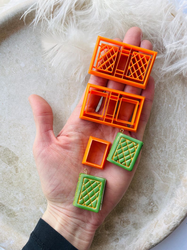 May include: Four orange plastic moulds for making polymer clay book charms. Two of the moulds are rectangular and have a grid pattern. The other two moulds are rectangular and have a simple frame design. Two green polymer clay book charms with a gold grid pattern are also shown.