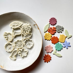 May include: A set of white plastic cookie cutters shaped like flowers and leaves, arranged on a white plate. Next to the plate are colorful fondant flowers and leaves.