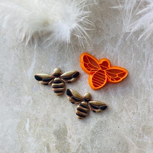 May include: Two gold bee-shaped cookie cutters with black stripes and an orange bee-shaped cookie cutter with a honeycomb pattern.