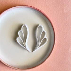 Puede incluir: Dos cortadores de galletas blancos con forma floral abstracta se muestran en un plato color crema con un borde rojo-marrón. El plato está sobre una superficie rosa claro. Los cortadores tienen un diseño curvo y en capas.