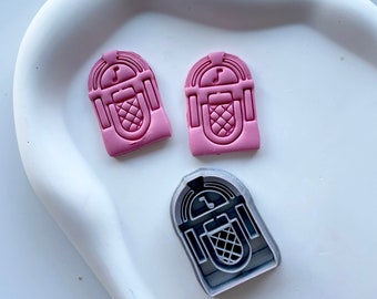 May include: Two pink and one gray plastic cookie cutters shaped like a vintage jukebox. The cookie cutters are on a white surface.