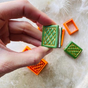 May include: A set of miniature book charms with a green cover and gold accents. The charms are made of polymer clay and have a detailed design. The charms are perfect for adding a touch of whimsy to your jewellery or crafts.