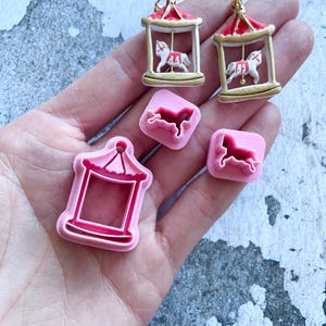 Puede incluir: Una colección de joyas y herramientas de manualidades con temática de carrusel. Dos pendientes de carrusel dorados con tejados rojos y caballos blancos. También hay cortadores de galletas rosas con forma de carrusel y un molde con forma de carrusel. Los artículos se muestran sobre un fondo texturizado.