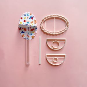 May include: A set of white plastic cookie cutters in the shape of an oval, a half-circle, and a smaller half-circle. The cutters are on a pink surface. There is also a clear glass test tube and a white plastic stick.
