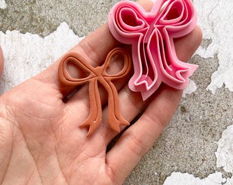 May include: Two bow-shaped cookie cutters. One is a light pink colour, and the other is a brown colour. The pink cutter has multiple layers, while the brown cutter is a single piece. The cutters are held in a hand.