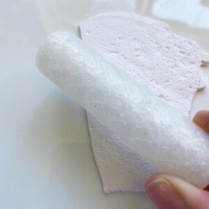 May include: A close-up shot shows a white, cylindrical object rolling over a light pink, textured surface. The object appears to be made of a porous material. The background is a plain white surface.