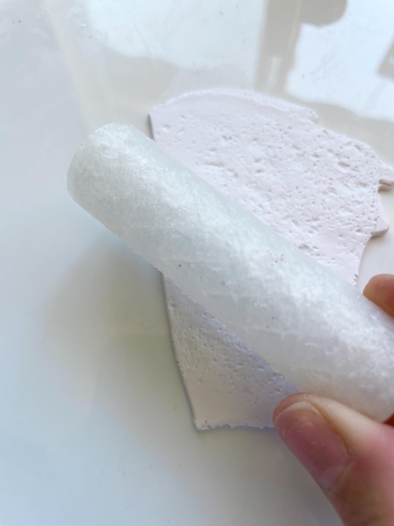 May include: A close-up shot shows a white, cylindrical object rolling over a light pink, textured surface. The object appears to be made of a porous material. The background is a plain white surface.