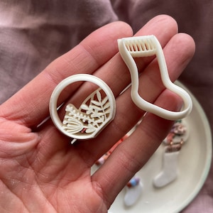 May include: Two white plastic cookie cutters, one shaped like a stocking and the other shaped like a leaf with a bird inside.
