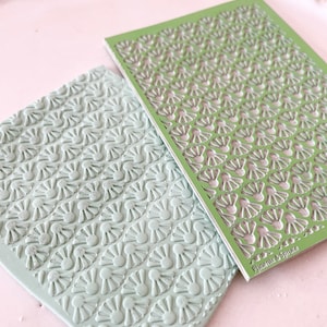 May include: A green plastic stencil with a repeating pattern of small flower shapes. The stencil is placed on top of a light green silicone mat with the same pattern embossed into the surface. The text "Phoenix & Spruce" is printed on the stencil.