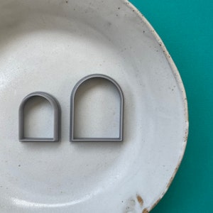 May include: Two grey, arch-shaped polymer clay cutters, one small and one large, sit on a white ceramic plate. The cutters have a simple, rounded top design. The plate has a speckled, rustic appearance, set against a teal background.