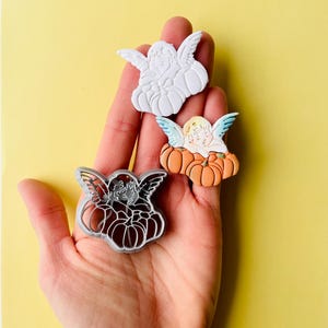 Puede incluir: Tres cortadores de galletas con forma de ángel sosteniendo una calabaza. Los cortadores son blancos, naranja y grises.