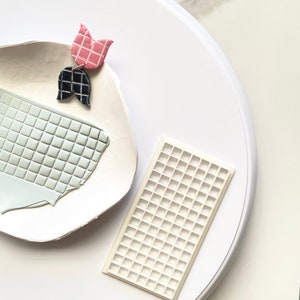 May include: A white ceramic plate with a light blue and white tiled design. A pink and black clay flower is on the plate. A white silicone mold with a grid pattern is on a white surface.