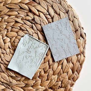 May include: Two light gray rectangular objects with botanical designs rest on a woven, brown surface. One object has raised leaf and stem patterns, while the other has a black line drawing of leaves. The objects are likely for crafting or design.