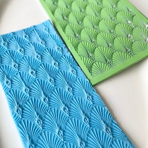 Puede incluir: Dos tapetes de hornear de silicona con un patrón decorativo de formas de abanico superpuestas. El tapete superior es verde y el tapete inferior es azul.