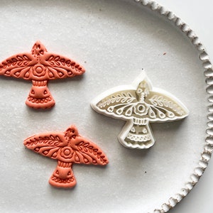 May include: Two terracotta-coloured bird-shaped cookie cutters and a white bird-shaped cookie cutter on a white ceramic plate with a scalloped edge.