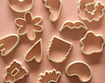May include: A set of 11 white plastic cookie cutters in various shapes, including a flower, a heart, a mustache, a sun, a cloud, and a crescent moon. The cutters are arranged on a light pink background.