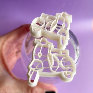 K&ouml;nnte beinhalten: Zwei wei&szlig;e Plastik-Ausstecher in Form von Vintage-Rollern. Die Ausstecher werden in einer Hand vor einem violetten Hintergrund gehalten.