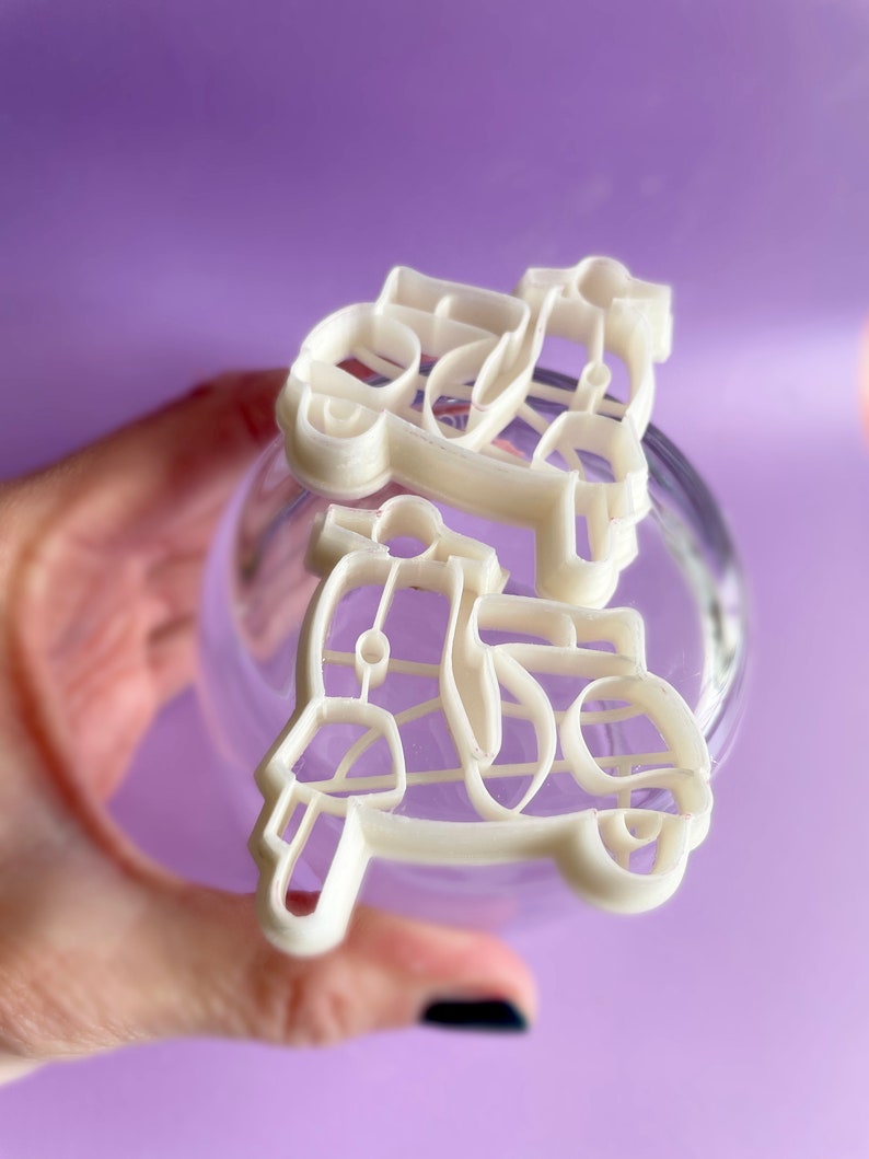 K&ouml;nnte beinhalten: Zwei wei&szlig;e Plastik-Ausstecher in Form von Vintage-Rollern. Die Ausstecher werden in einer Hand vor einem violetten Hintergrund gehalten.