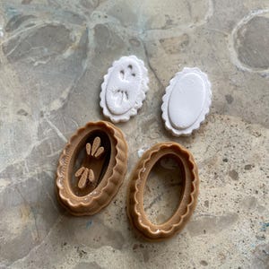 May include: Oval-shaped cookie cutters in brown and white. Two brown cutters have a scalloped edge, one with a floral design inside. Two white cutters have a scalloped edge, one with a floral design. The cutters are on a marble surface.