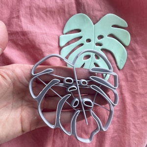 May include: A gray plastic cookie cutter in the shape of a monstera leaf. The cutter is being held in a hand.
