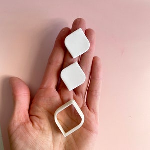 May include: Three white plastic cookie cutters in the shape of a diamond, a square, and a rounded rectangle. The cutters are held in a person's hand.