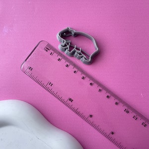May include: A gray plastic cookie cutter in the shape of a bear, approximately 3 inches long, sits on a pink surface. A clear ruler with both inches and centimeters is next to the cutter. The ruler is angled across the image.
