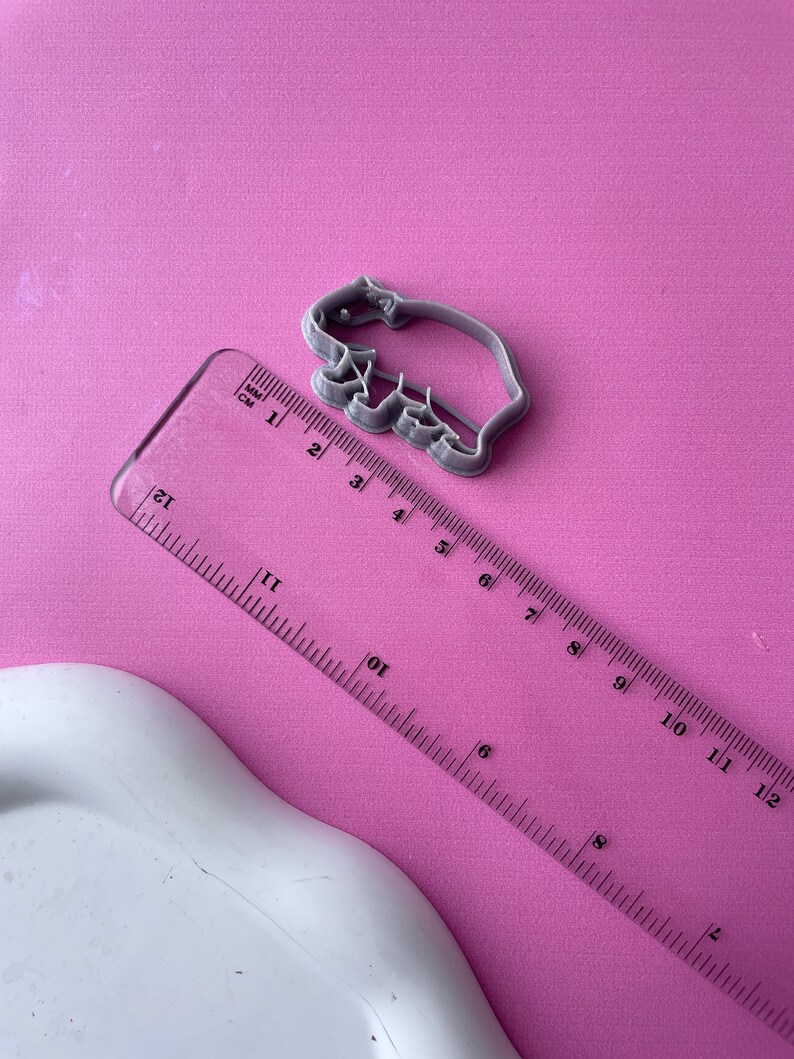 May include: A gray plastic cookie cutter in the shape of a bear, approximately 3 inches long, sits on a pink surface. A clear ruler with both inches and centimeters is next to the cutter. The ruler is angled across the image.