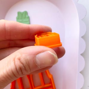 May include: Orange plastic cookie cutters shaped like a vest and a square. The vest cutter has a grid pattern and the square cutter has a raised design.