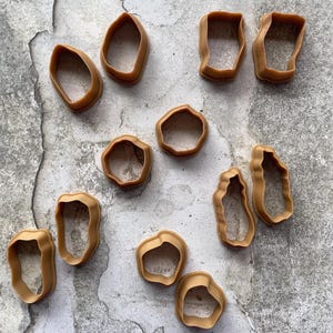 May include: A collection of tan-colored, irregularly shaped cookie cutters. The cutters are made of plastic and have a smooth, matte finish. The shapes vary, including ovals, rectangles, and organic forms. They are arranged on a textured, gray surface.