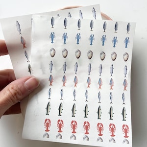 Op de afbeelding: Vellen stickers in aquarel-stijl met zeevruchten met verschillende vissen, blauwe kreeften en bruine oesterschelpen. De stickers zijn gerangschikt in nette rijen en kolommen, met een verscheidenheid aan kleuren.