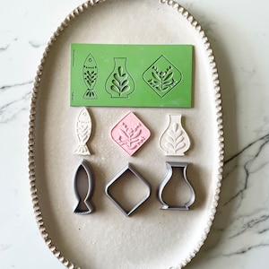 Pode incluir: Um conjunto de seis cortadores de biscoitos em várias formas, incluindo um peixe, um vaso e um diamante. Os cortadores são feitos de plástico cinza claro e estão dispostos em um prato oval branco. O prato tem uma borda decorativa.