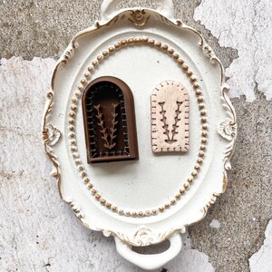 Puede incluir: Dos objetos decorativos en forma de arco con diseños florales descansan sobre una bandeja blanca y ornamentada. Uno es marrón oscuro, el otro es de color beige claro. La bandeja tiene detalles dorados y un borde de cuentas. El fondo es una superficie texturizada blanquecina.