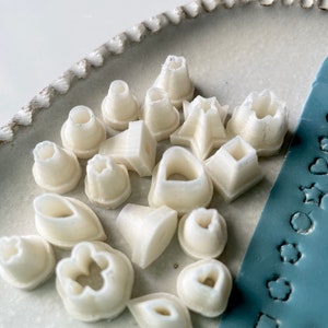 May include: A collection of 15 white plastic cookie cutters in various shapes, including stars, hearts, leaves, and flowers. The cutters are arranged on a white ceramic plate.