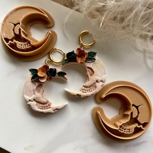 May include: Two crescent moon-shaped earrings featuring a skull design, adorned with small black and orange flowers and green leaves, attached to gold-toned hoops. Two tan plastic crescent moon-shaped cutters are positioned next to the earrings.