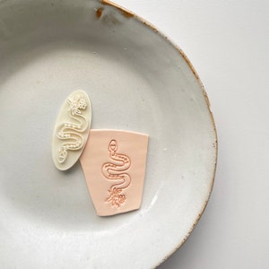 Könnte beinhalten: Zwei weiße Gummistempel mit einem Schlangen- und Blumendesign. Die Stempel befinden sich auf einem weißen Keramikteller.