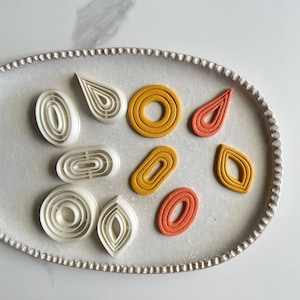 Könnte beinhalten: Ein Set mit neun Ausstechern in verschiedenen Formen, darunter Kreise, Ovale und Tropfenformen. Die Ausstecher sind aus weißem Kunststoff und auf einem weißen Teller angeordnet. Einige der Ausstecher sind mit buntem Teig gefüllt.