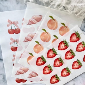 May include: Sheets of fruit-themed stickers featuring cherries with pink bows, seashells, peaches, and strawberries. The stickers are printed on white paper and arranged in a layered composition. The stickers have a watercolor style.