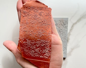 May include: A close-up of a hand holding a terracotta-coloured clay tile with a floral design embossed into the surface. The tile is slightly irregular in shape and has a rough texture.