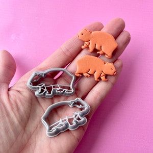 Könnte beinhalten: Zwei orangefarbene Capybara-Ausstecher und zwei orangefarbene Capybara-Kekse werden gezeigt. Die Ausstecher sind grau und aus Kunststoff. Die Kekse haben eine einfarbige orange Farbe. Die Artikel werden in einer Hand vor einem rosa Hintergrund gehalten.