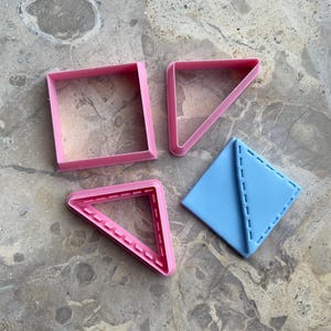 Puede incluir: Cuatro cortadores de galletas geométricos en rosa y azul. Las formas incluyen un cuadrado, dos triángulos y un rombo. Los cortadores son de plástico y están dispuestos sobre una superficie de mármol.